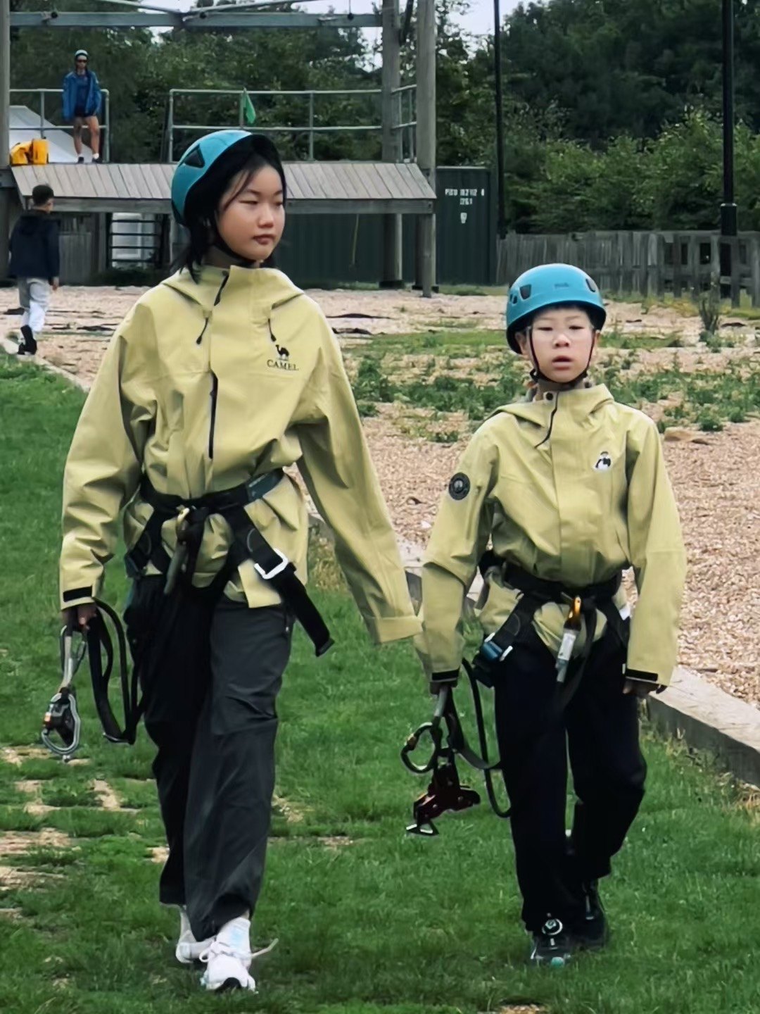 Two participants wearing helmets and harnesses walk across an outdoor activity area